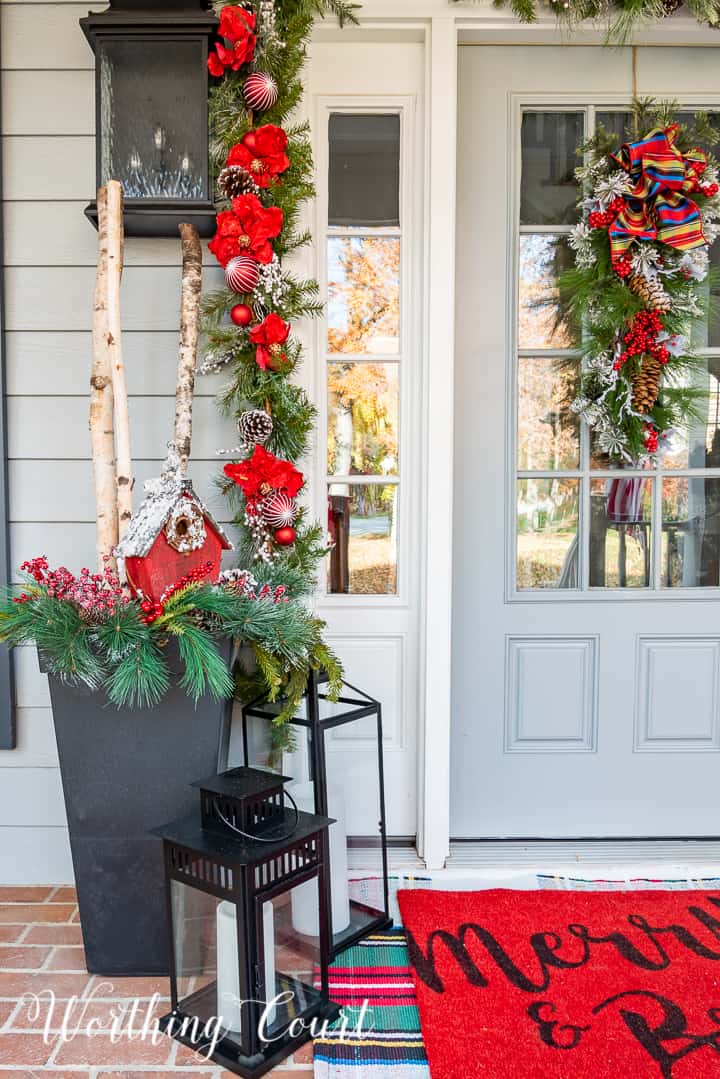 front porch decorated for Christmas with red and white garland, planters, trees, lanterns, rugs and pillows