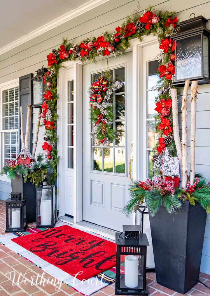 front porch decorated for Christmas with red and white garland, planters, trees, lanterns, rugs and pillows