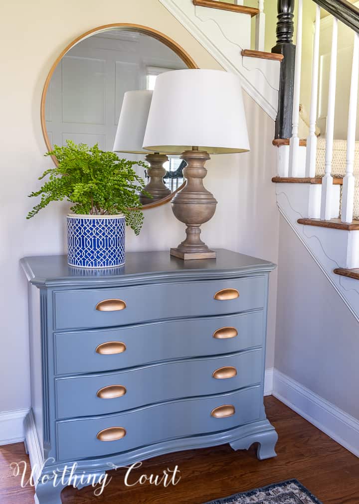 round mirror above a gray chest with lamp and planter