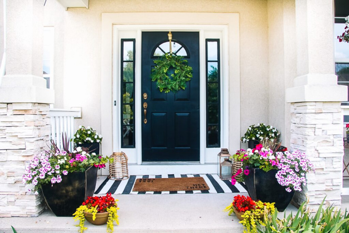 small front porch filled with pots of flowers on a white brick house with a black door