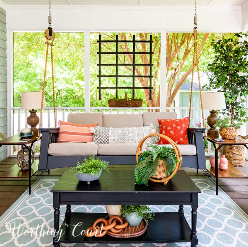 A screen porch decorated with a swing, end tables, lamps, artificial tree and chairs