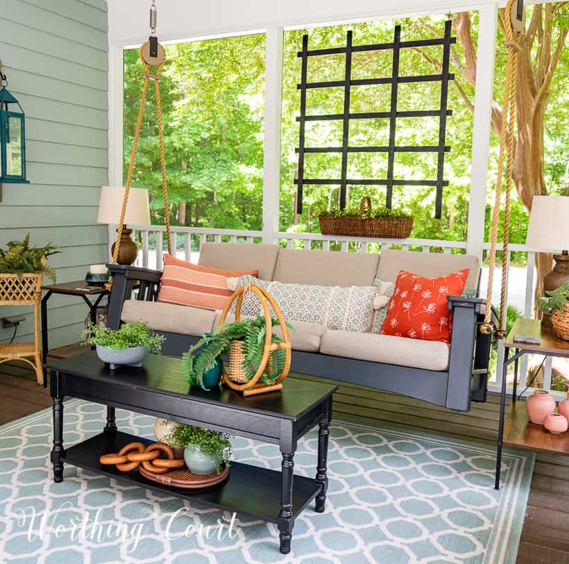 A screen porch decorated with a swing, end tables, lamps, artificial tree and chairs