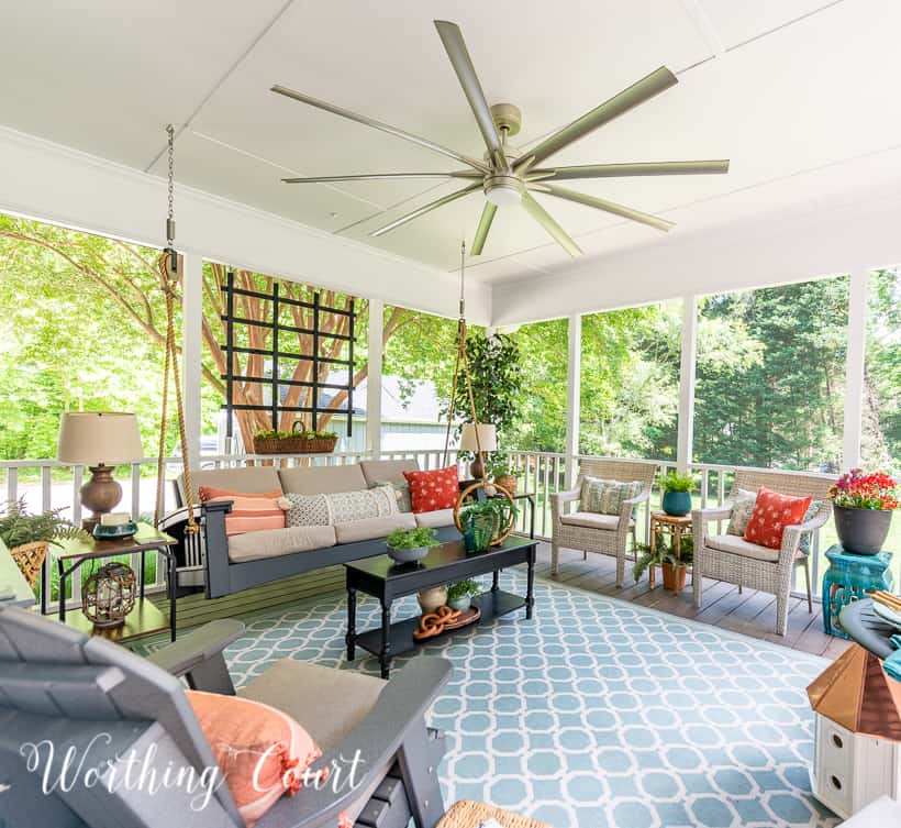 A screen porch decorated with a swing, end tables, lamps, artificial tree and chairs