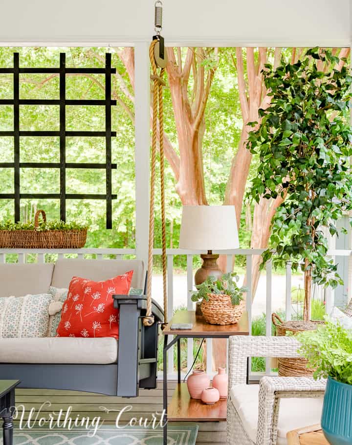 A screen porch decorated with a swing, end tables, lamps, artificial tree and chairs