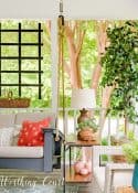 A screen porch decorated with a swing, end tables, lamps, artificial tree and chairs