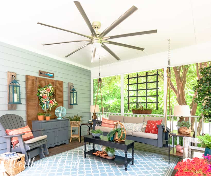 A screen porch decorated with a swing, end tables, lamps, artificial tree and chairs