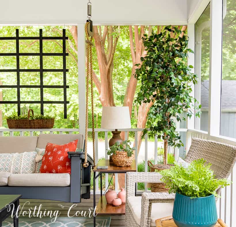 A screen porch decorated with a swing, end tables, lamps, artificial tree and chairs