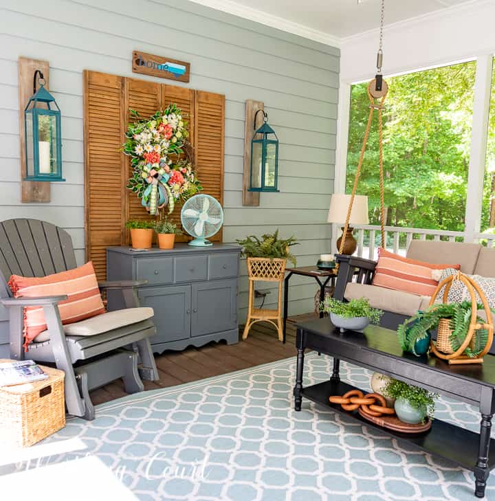 A screen porch decorated with a swing, end tables, lamps, artificial tree and chairs