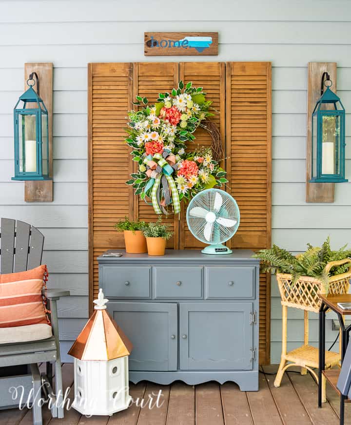 Gray chest with a wreath hanging above it on old bifold doors, flanked by a glider and a plant stand