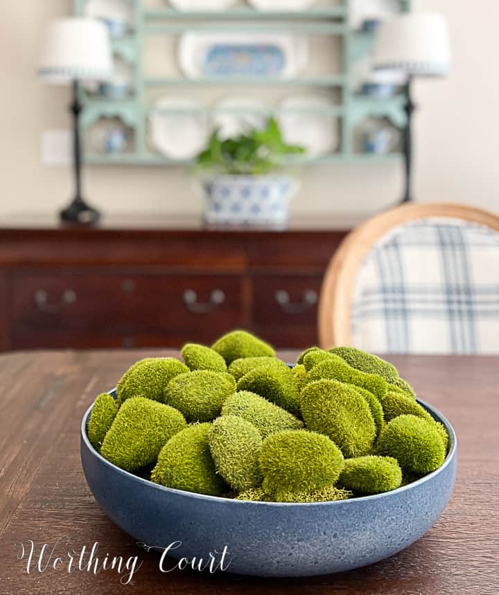 blue bowl filled with artificial moss covered rocks on a dining table with a plate rack in the background