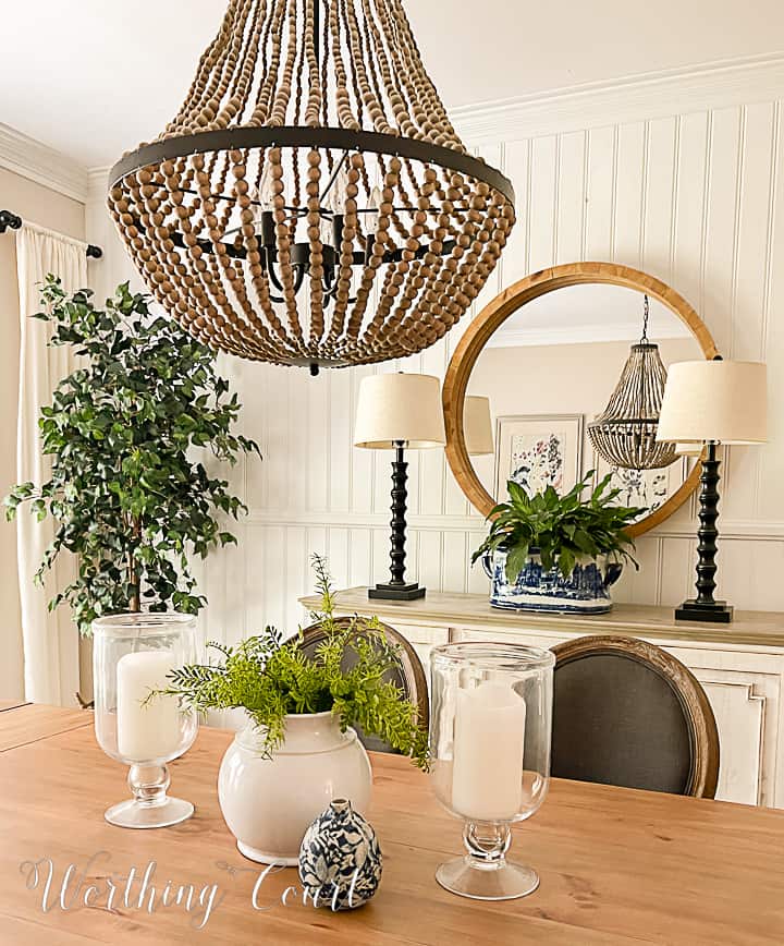 Summer decorations in a dining room consisting of a pretty blue and white urn on the sideboard and a faux plant with large glass hurricane lanterns for the table centerpiece. The type of urn on the sideboard is actually called a footbath and looks just gorgeous filled with a large peace lily