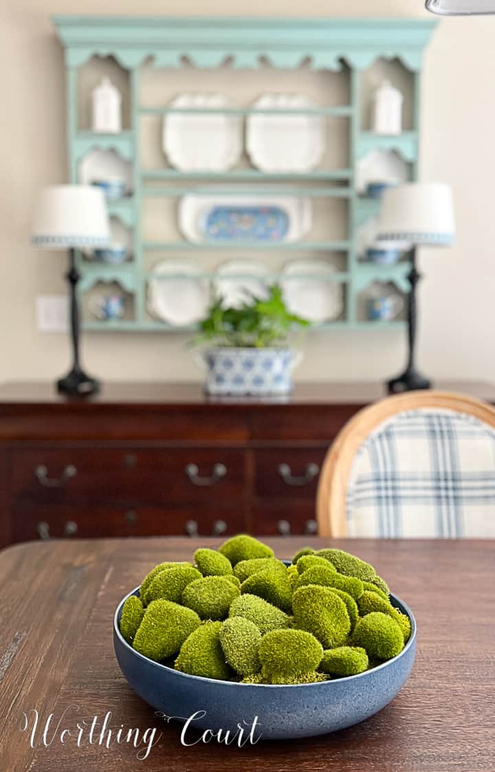 blue bowl filled with artificial moss covered rocks on a dining table with a plate rack in the background