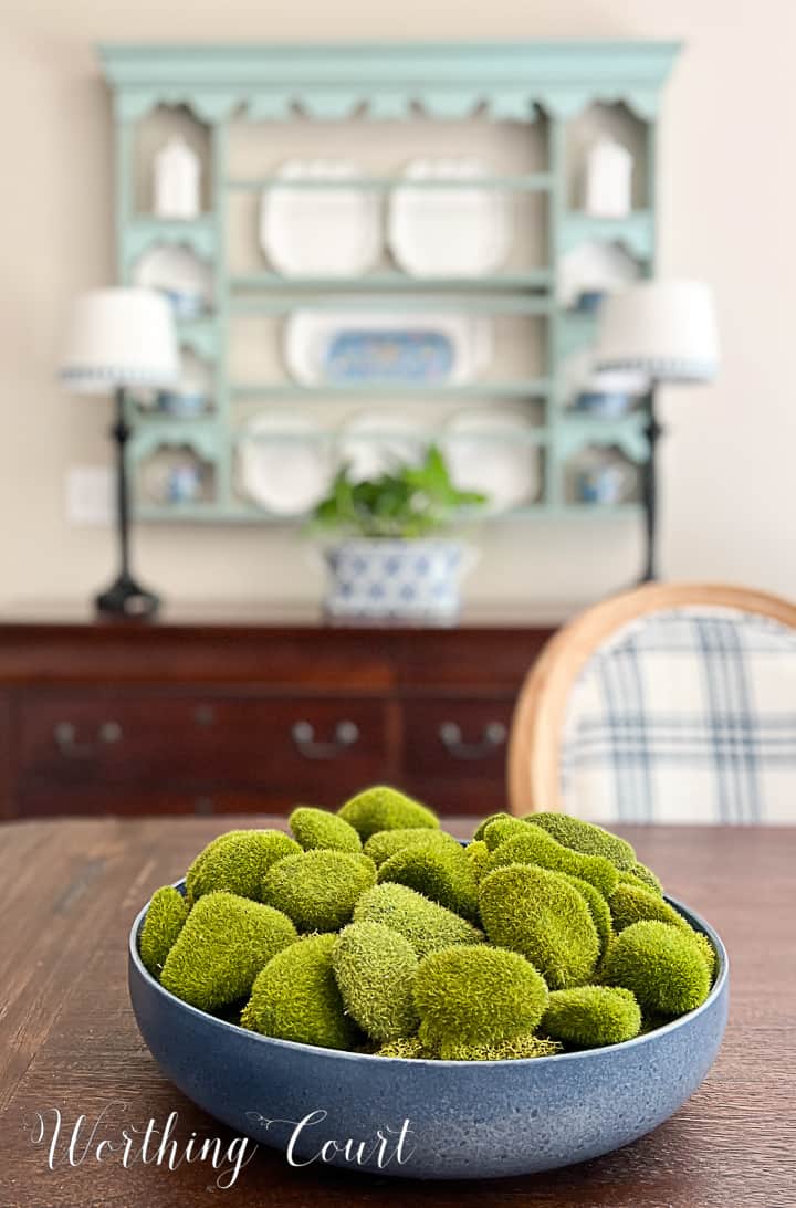 blue bowl filled with artificial moss covered rocks on a dining table with a plate rack in the background