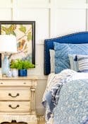 partial image of bed with blue headboard and blue and white bedding and night stand with art