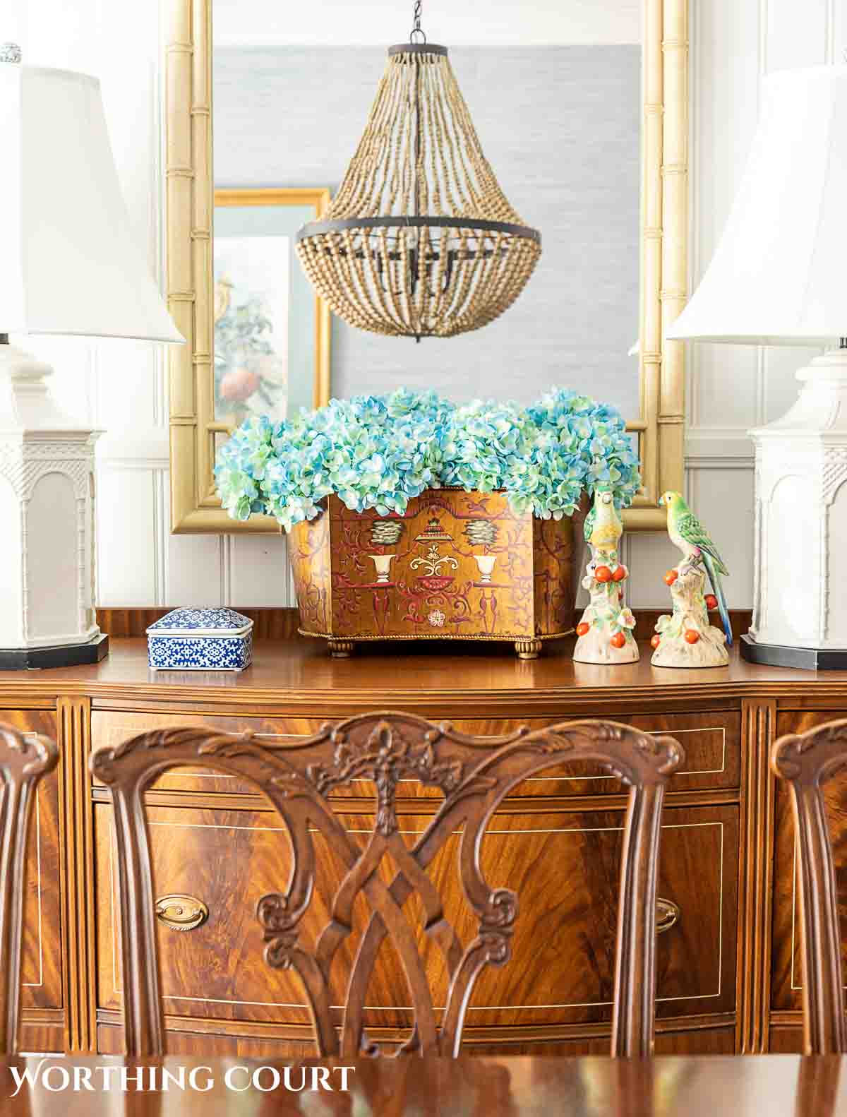 faux blue hydrangeas in an oblong container on a sideboard in front of a mirror flanked by white lamps