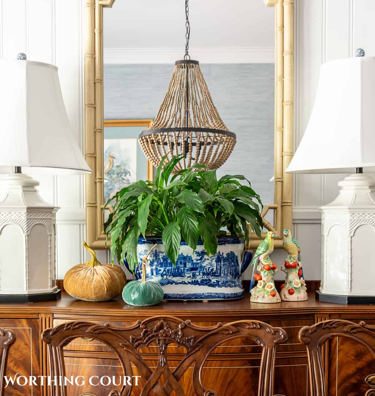 faux bamboo mirror hanging above a wood sideboard with two white lamps and fall decorations