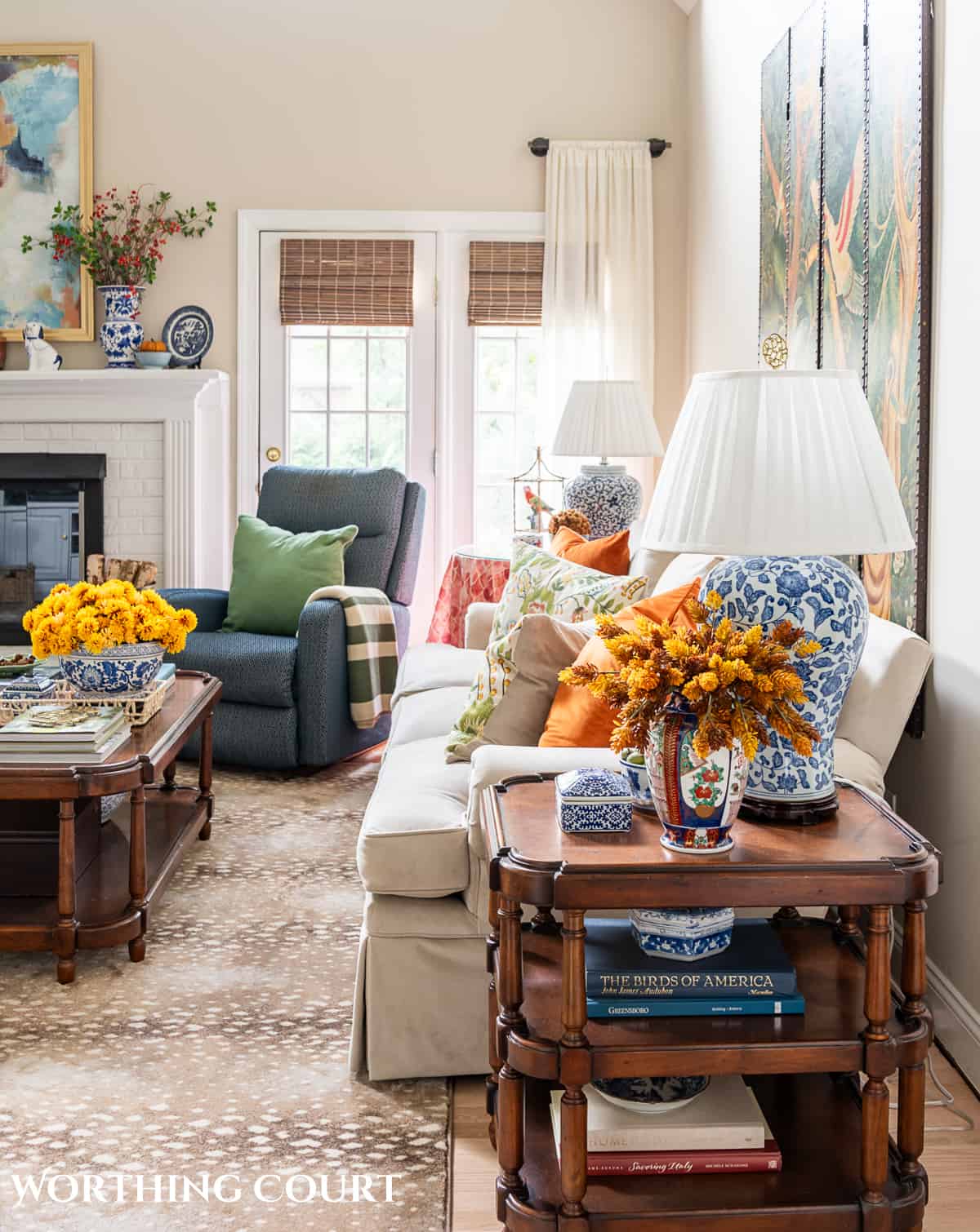 side view of beige sofa and blue chair beside white brick fireplace with fall decorations and a blue and white lamp on a brown wood end table