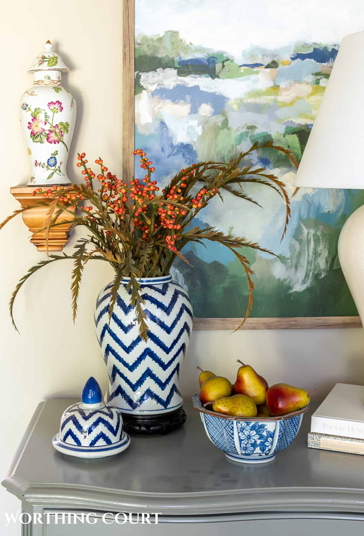 blue and white ginger jar with faux fall stems and a bowl of fake pears beside a white lamp on a gray chest