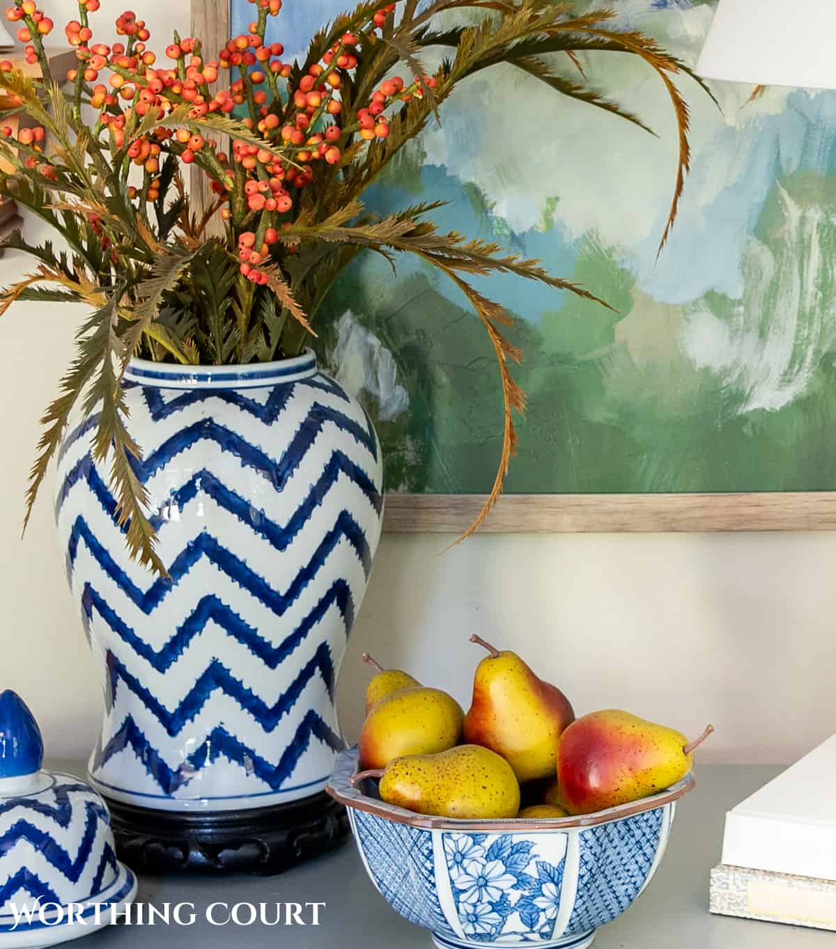 blue and white ginger jar with faux fall stems and a bowl of fake pears beside a white lamp on a gray chest