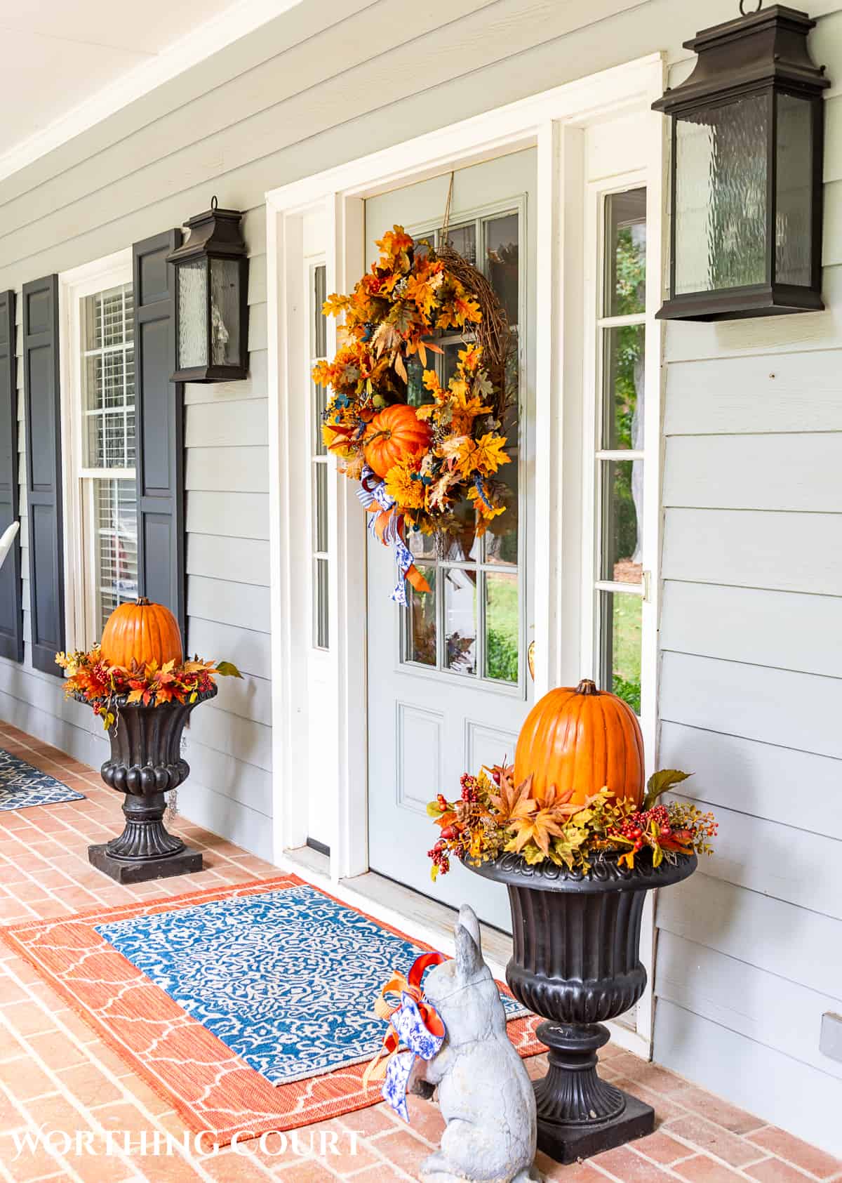 Gray front door on a large front porch with fall wreath flanked by two planters with fall decorations and large pumpkins