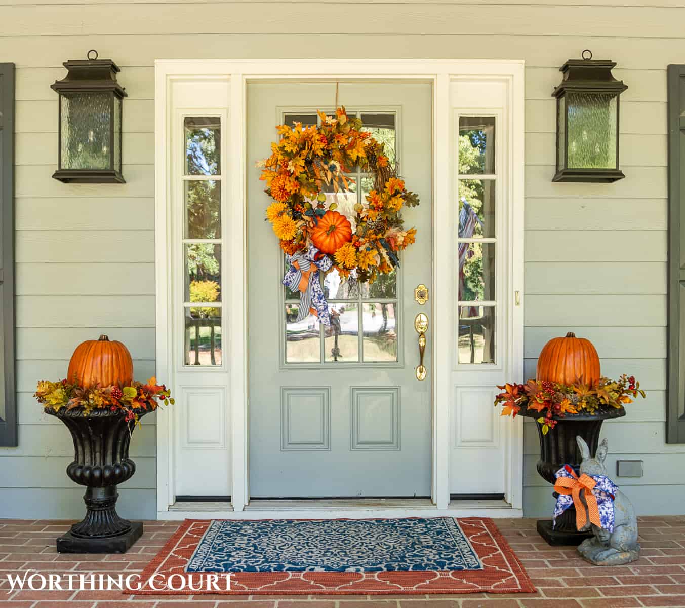Gray front door on a large front porch with fall wreath flanked by two planters with fall decorations and large pumpkins