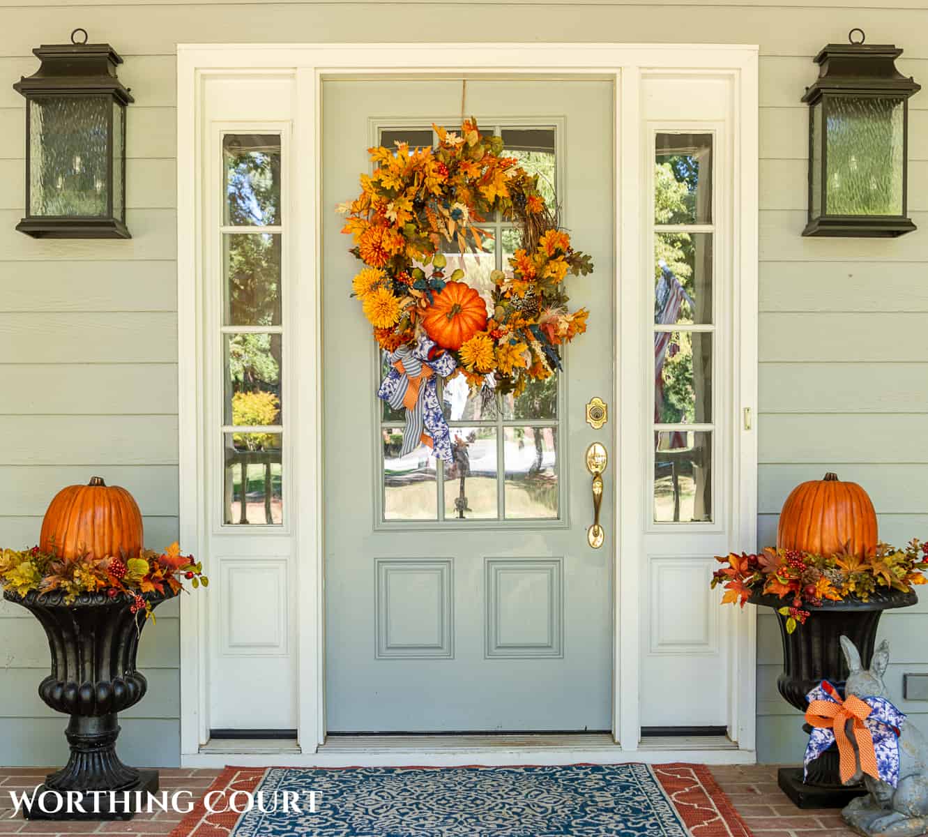 Gray front door on a large front porch with fall wreath flanked by two planters with fall decorations and large pumpkins