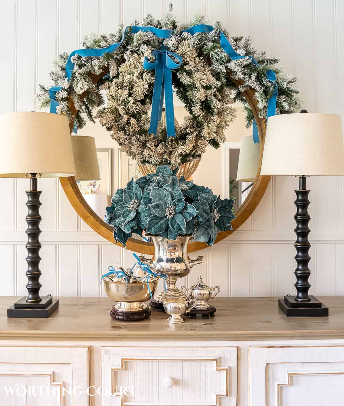 blue faux poinsettias in a silver urn in front of a mirror on a sideboard flanked by a pair of tall lamps