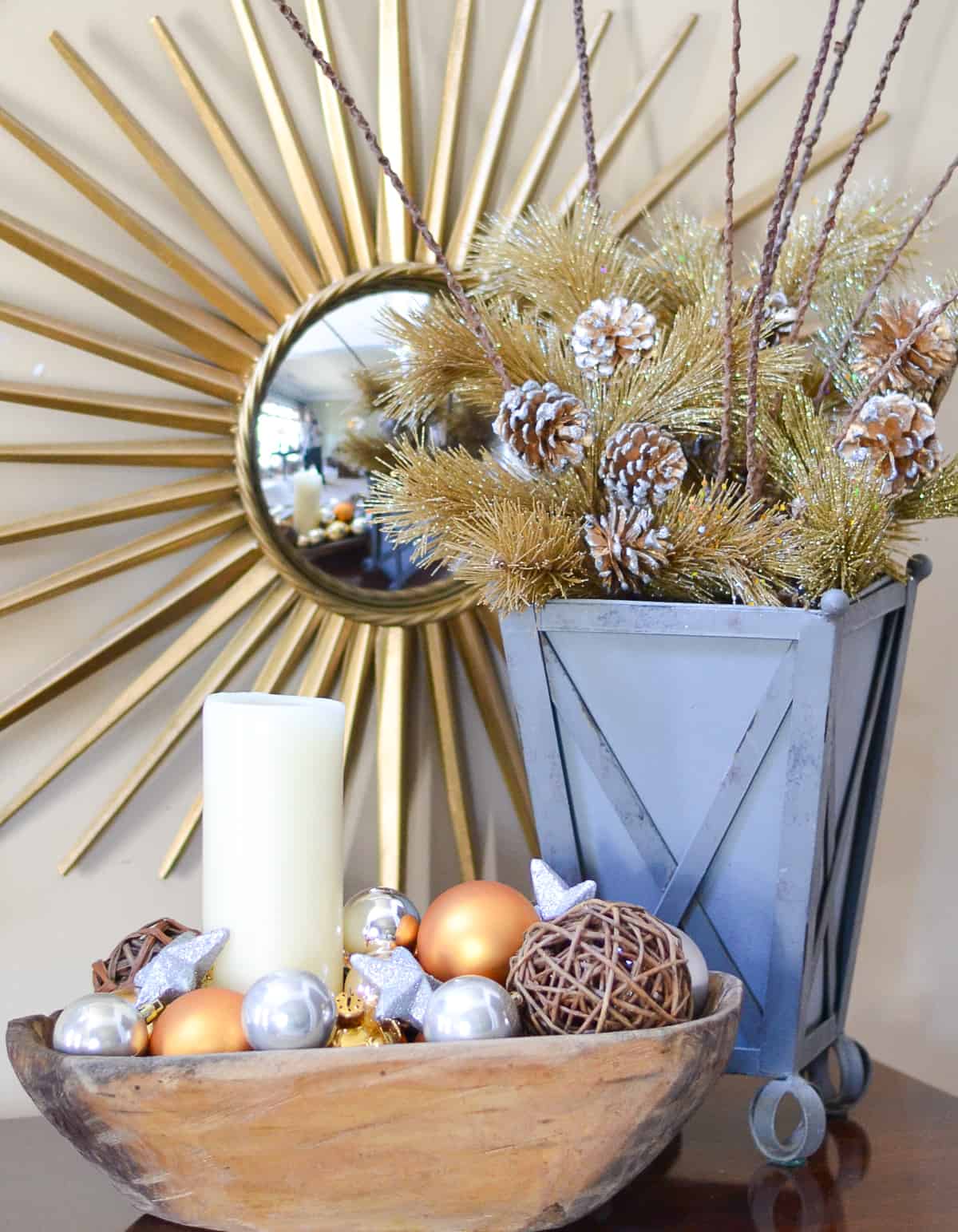 gold and silver ornaments and a wood bowl and a gold and pine cone arrangement in a gray container in front of a sunburst mirror