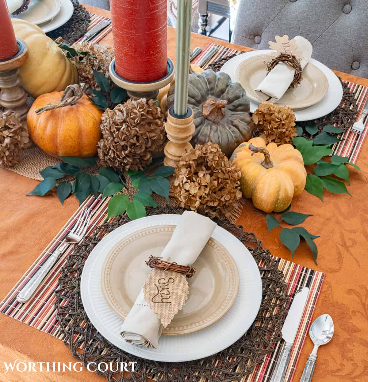 Table set for Thanksgiving with a rust colored tablecloth and candles, white dishes and a centerpiece made with pumpkins, foliage and dried hydrangeas