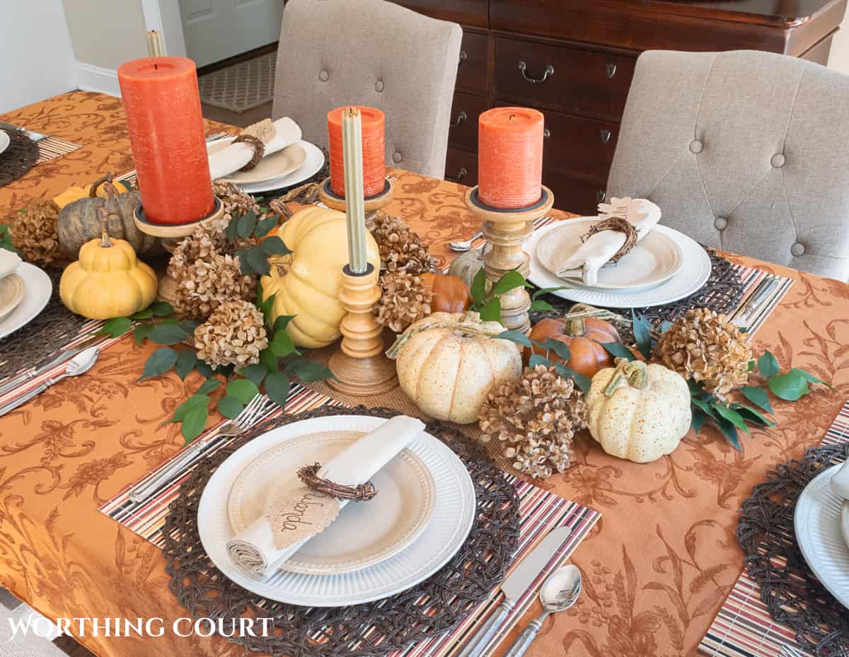 Table set for Thanksgiving with a rust colored tablecloth and candles, white dishes and a centerpiece made with pumpkins, foliage and dried hydrangeas
