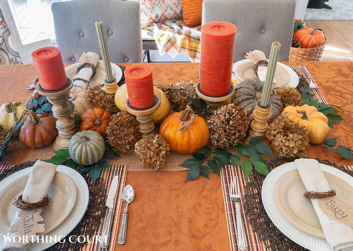 Table set for Thanksgiving with a rust colored tablecloth and candles, white dishes and a centerpiece made with pumpkins, foliage and dried hydrangeas