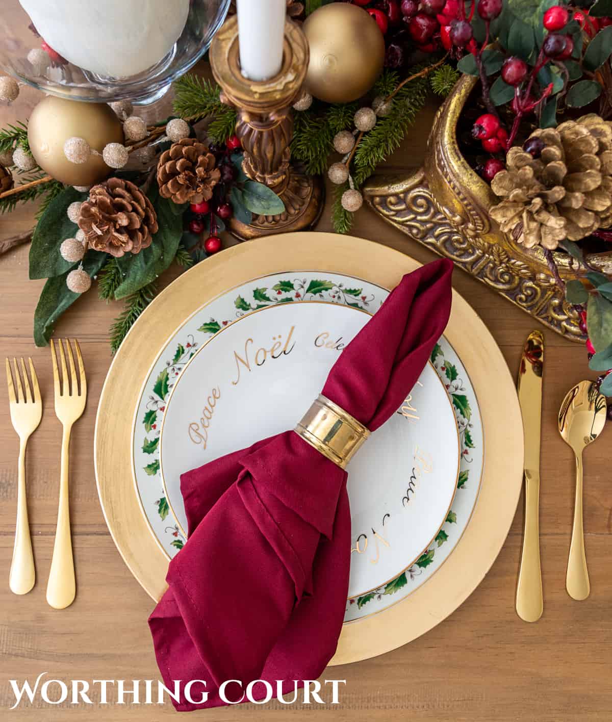 Christmas place setting with white plates stacked on a gold charger, with gold siverware and a burgundy napkin
