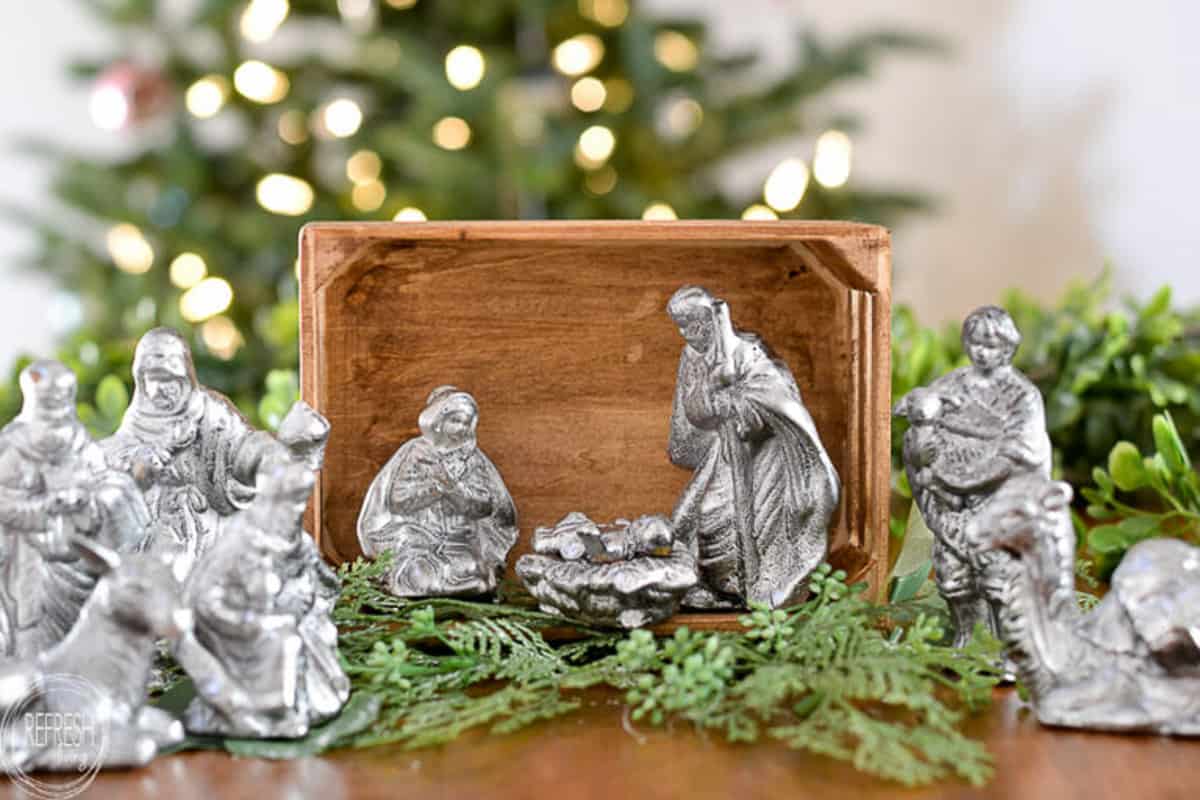 nativity scene display with silver figurines in a wood box surrounded by faux greenery