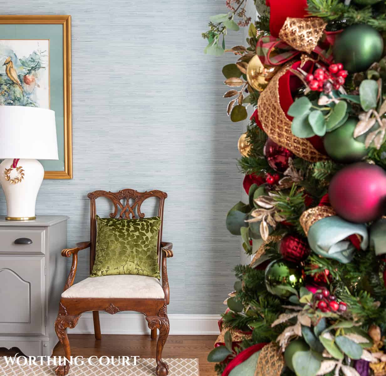 Christmas tree with green, gold and burgundy decorations in the foreground with a chair and pillow in the background