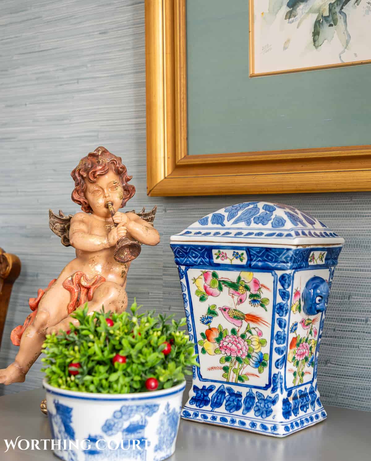 Vignette on a gray chest with a vintage cherub, blue and white urn and bowl with a boxwood orb with red berries