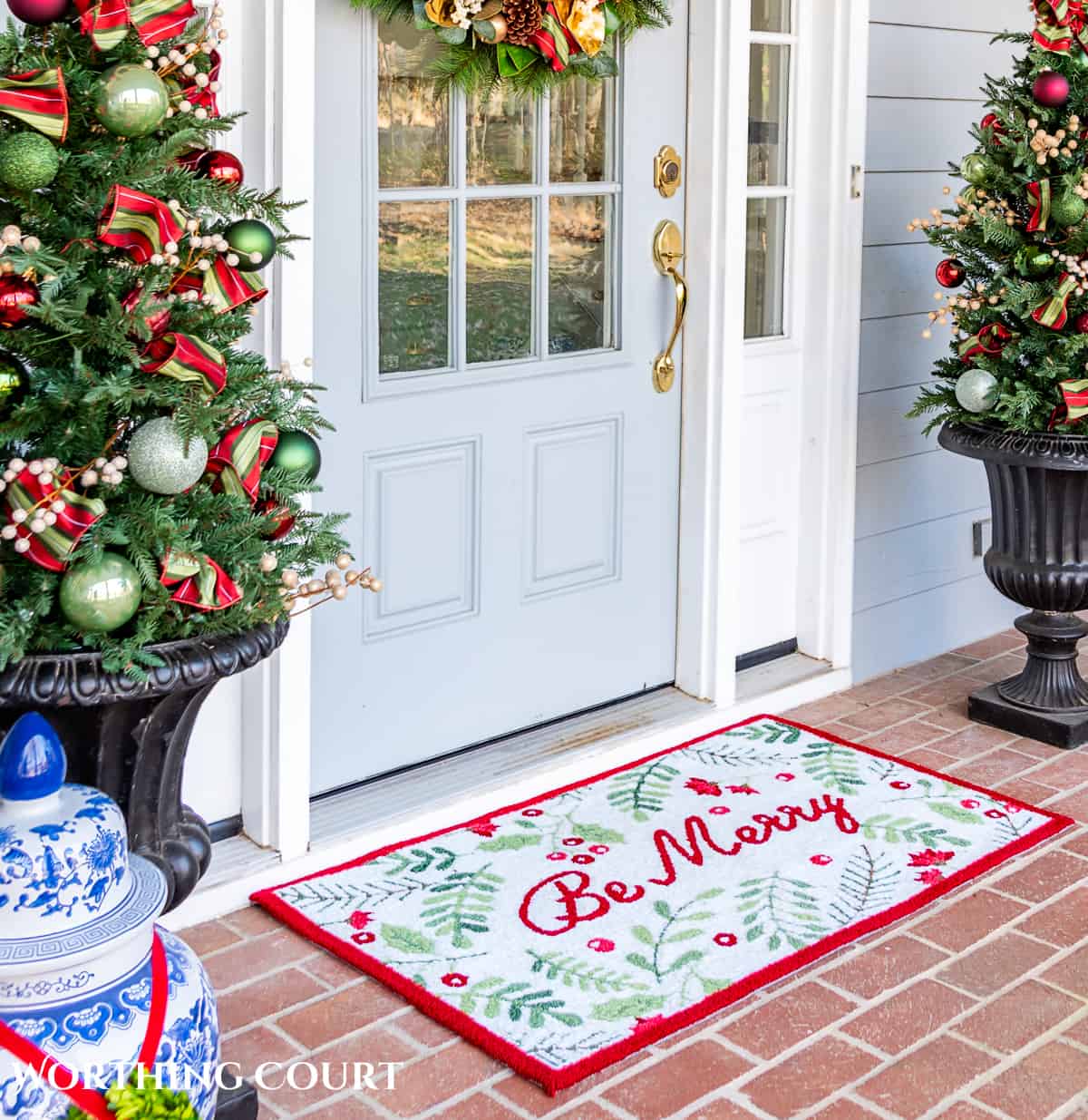 red, green and white Christmas welcome mat with the words "Be Merry" in front of a gray front door flanked by small Christmas trees in urns