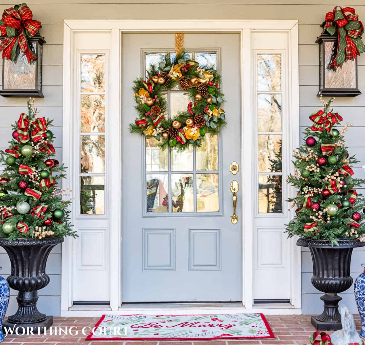 front door with a Christmas wreath flanked by urns with small trees with red, green and gold Christmas decorations on them