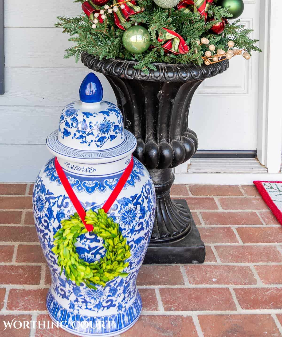 large blue and white ginger jar with a boxwood wreath around it in front of a  planter on a front porch decorated for Christmas