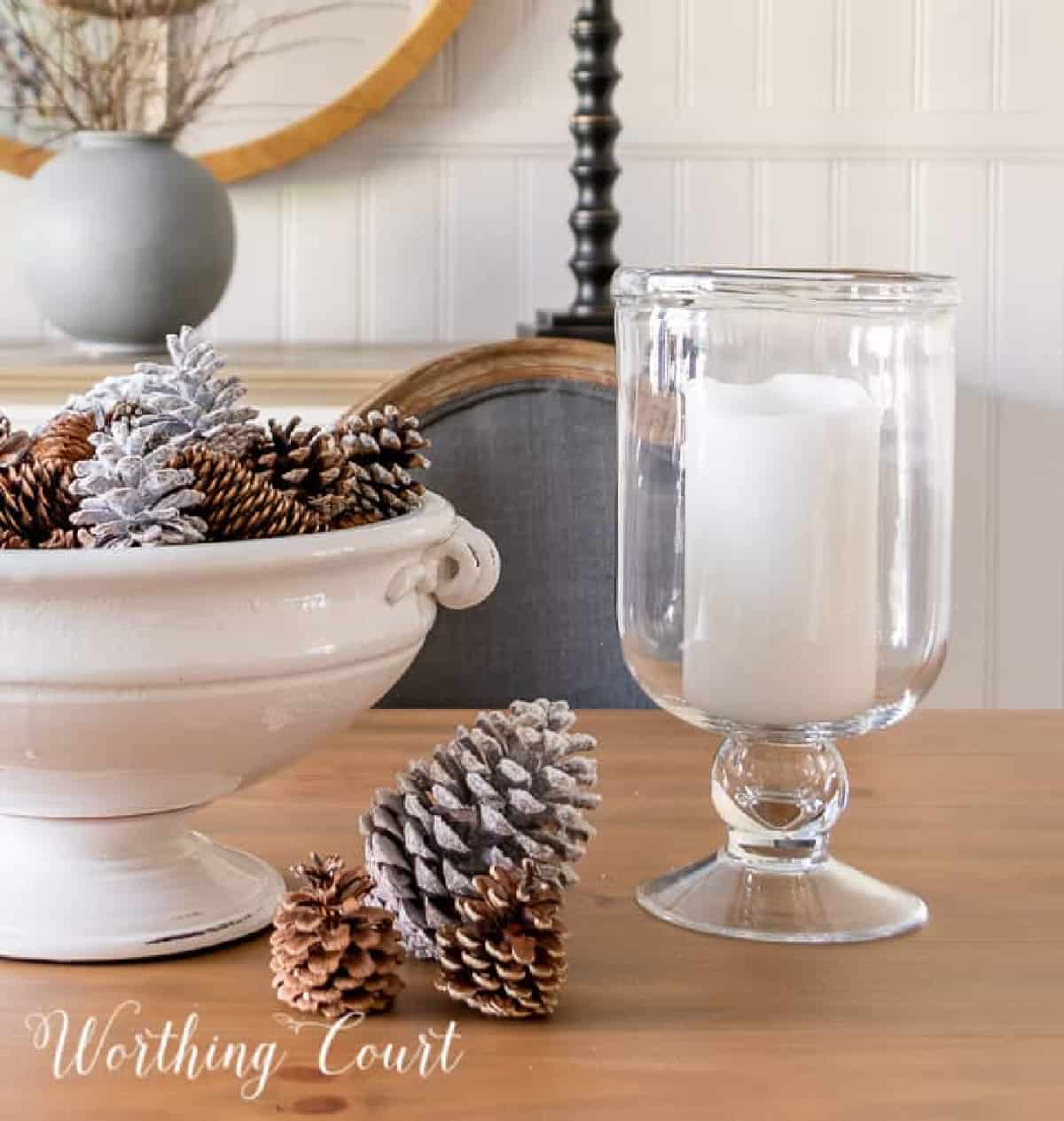 large white pedestal bowl on a dining table decorated for winter with pinecones and flanked by glass hurricanes
