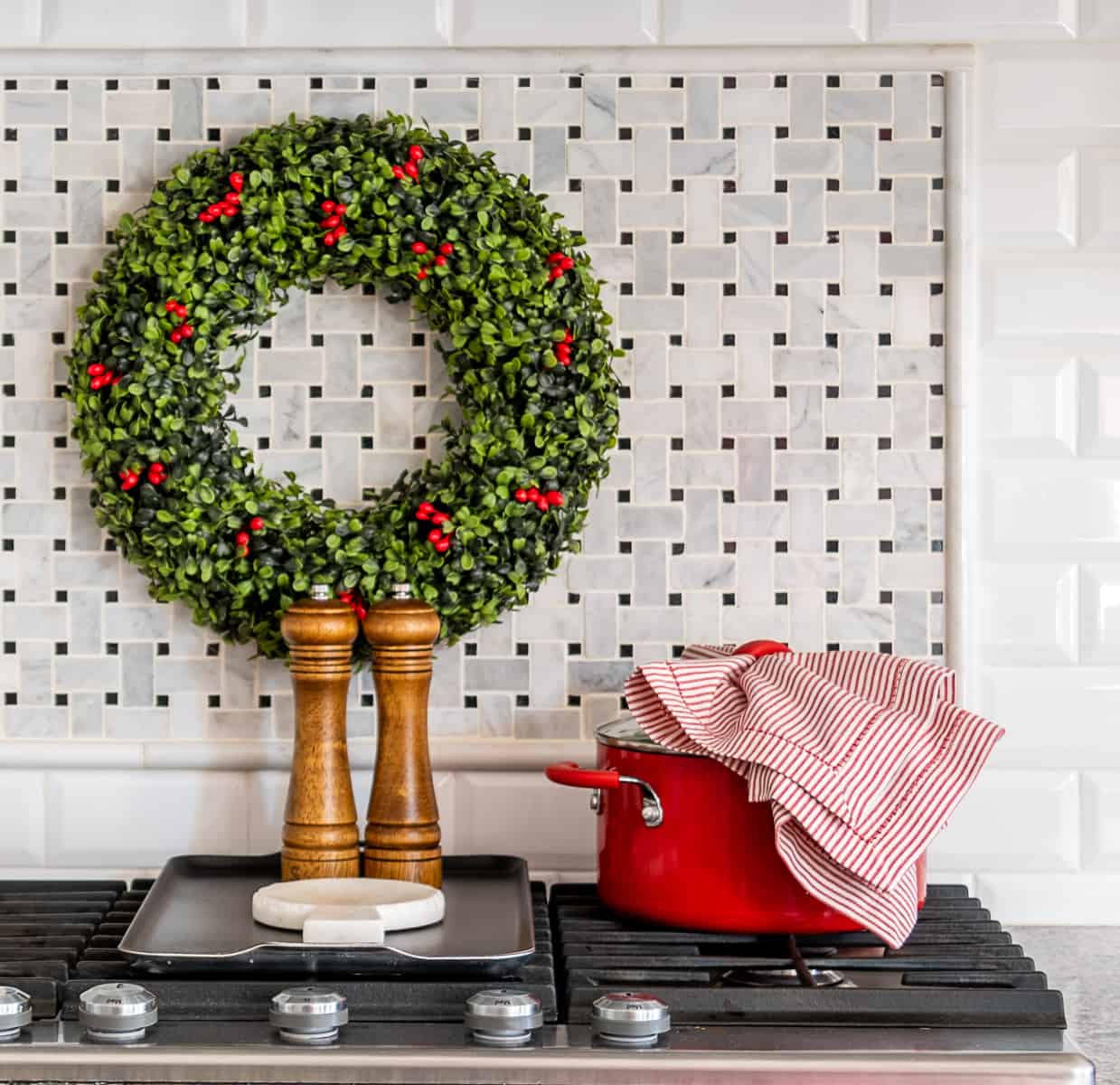 Christmas wreath on backsplash above cooktop with red dutch oven, red and white striped kitchen towel and wood salt and pepper mills