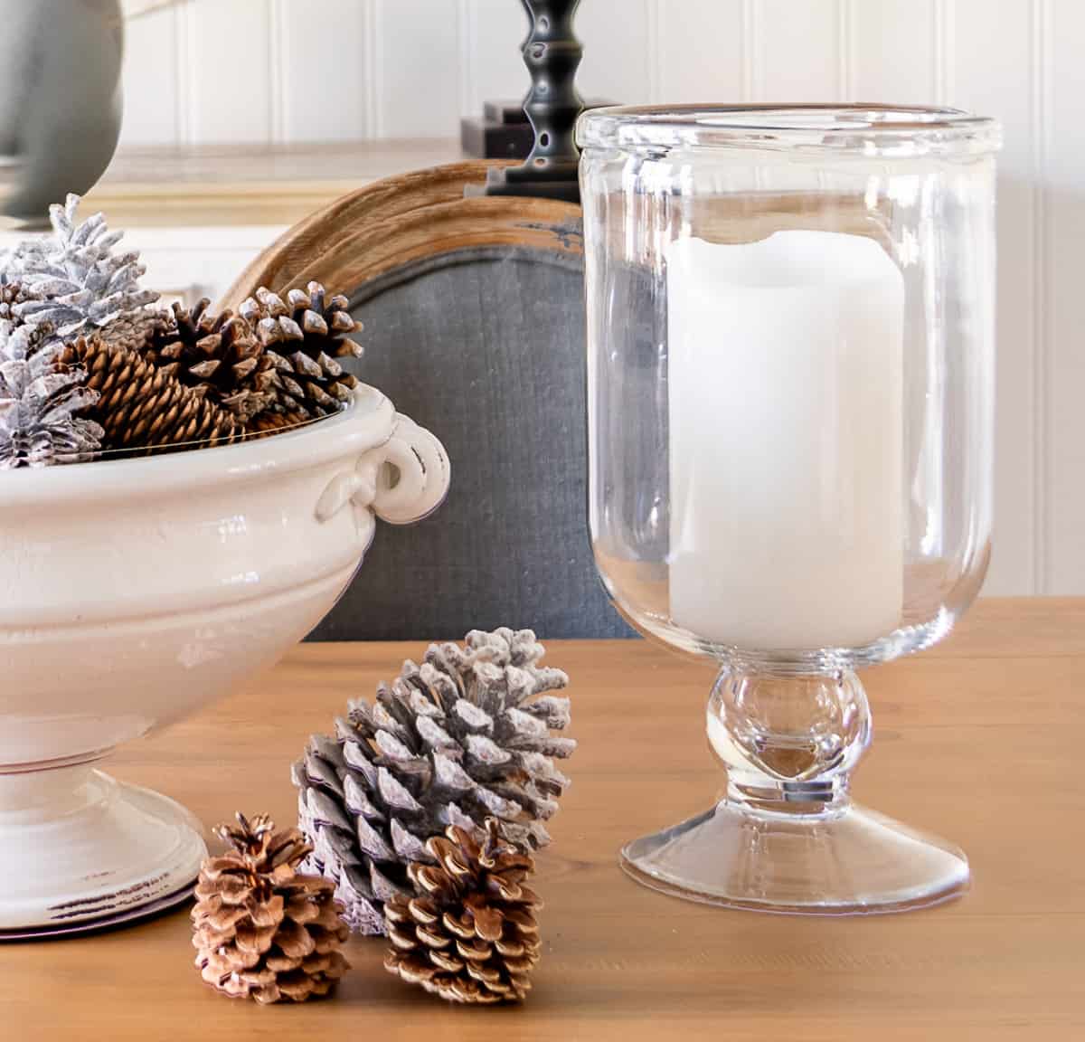 large white pedestal bowl on a dining table decorated for winter with pinecones and flanked by glass hurricanes