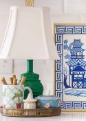 round brass tray on a kitchen counter holding a small green lamp, a mug with spoons and a sugar bowl in front of a white subway tile backsplash