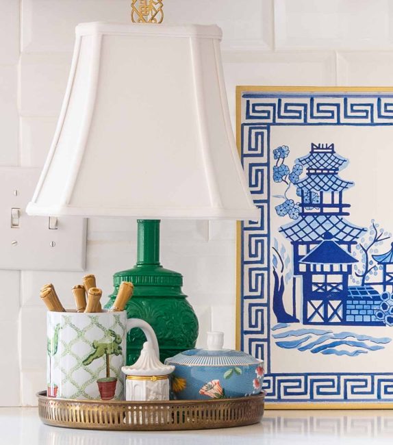 round brass tray on a kitchen counter holding a small green lamp, a mug with spoons and a sugar bowl in front of a white subway tile backsplash