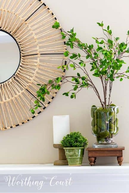 vignette of natural looking faux greenery branches in a glass vase, a candle, and a small planter on a mantel