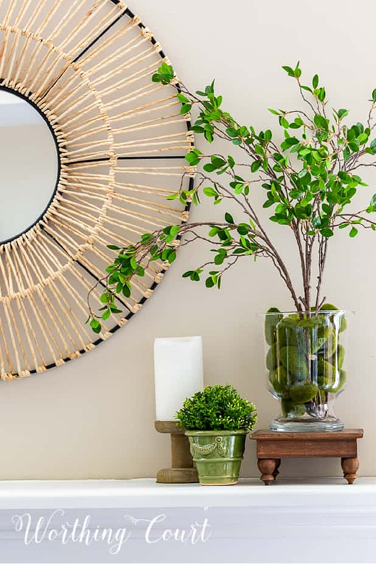 vignette of natural looking faux greenery branches in a glass vase, a candle, and a small planter on a mantel
