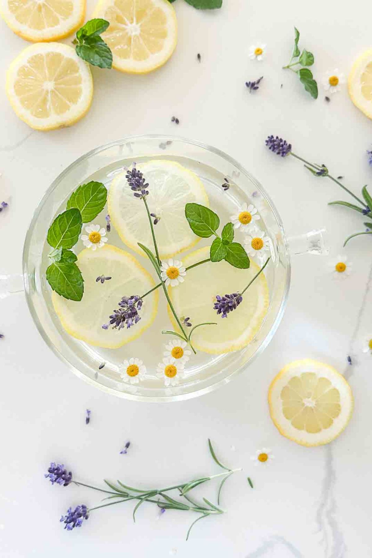 a clear glass simmer pot filled with clear liquid with daisies, leaves, and purple flowers floating in it
