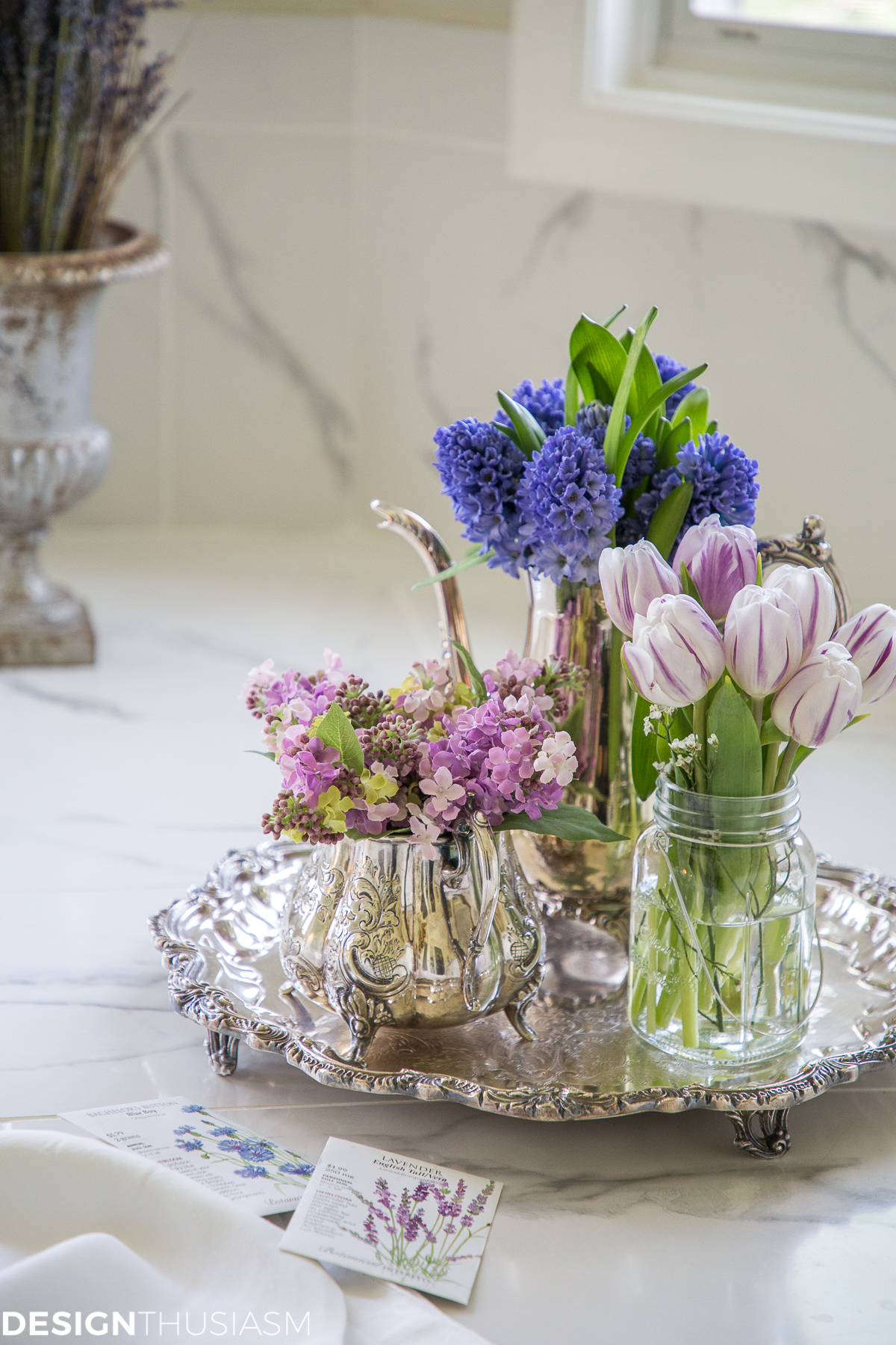round footed silver tray on a marble bathroom counter holding three vessels filled with lavender colored flowers