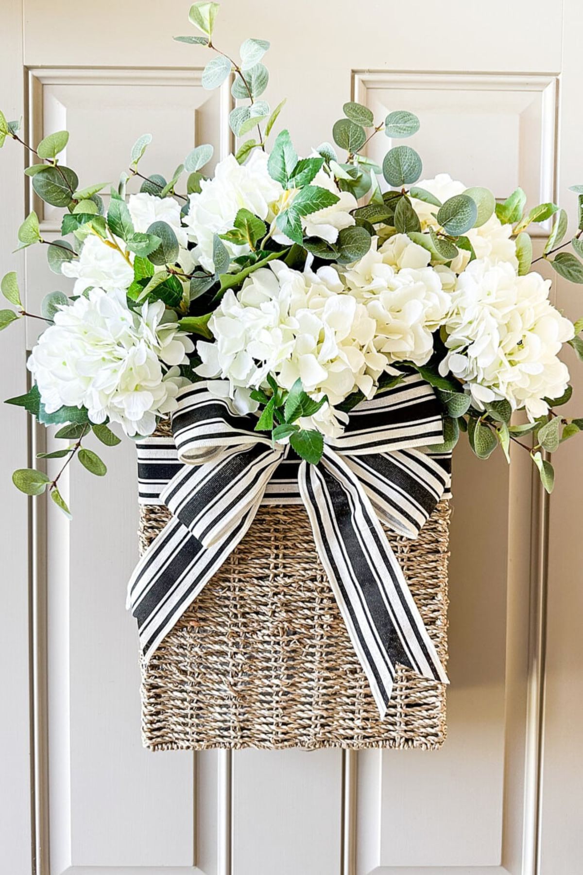wicker basket hanging on a beige front door filled with faux white geraniums and embellished with a black and white striped bow