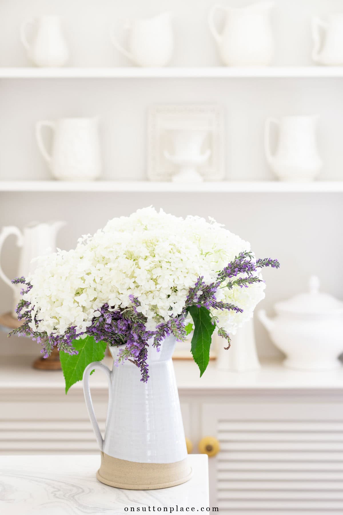 white cut flowers in a white vase in front of shelves containing white dishes