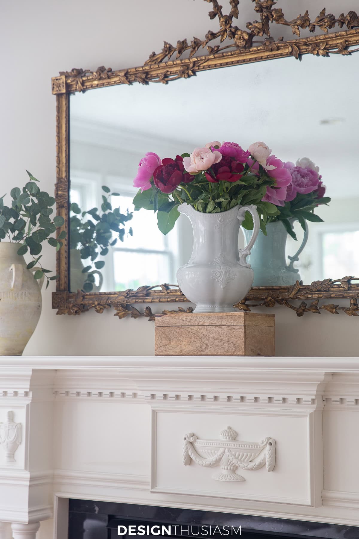 white pitcher filled with various shades of pink flowers in front of a mirror on a mantel